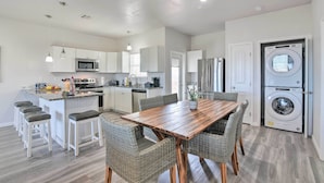 A modern kitchen with white cabinets, a central island with stools, a dining table with wicker chairs, and a built-in washer and dryer. - Near Ocean: Surfside Beach Gem w/ Deck & Views! (Freeport)