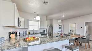 A modern kitchen with white cabinets, a marble countertop, and a dining area with a wooden table and wicker chairs. - Near Ocean: Surfside Beach Gem w/ Deck & Views! (Freeport)
