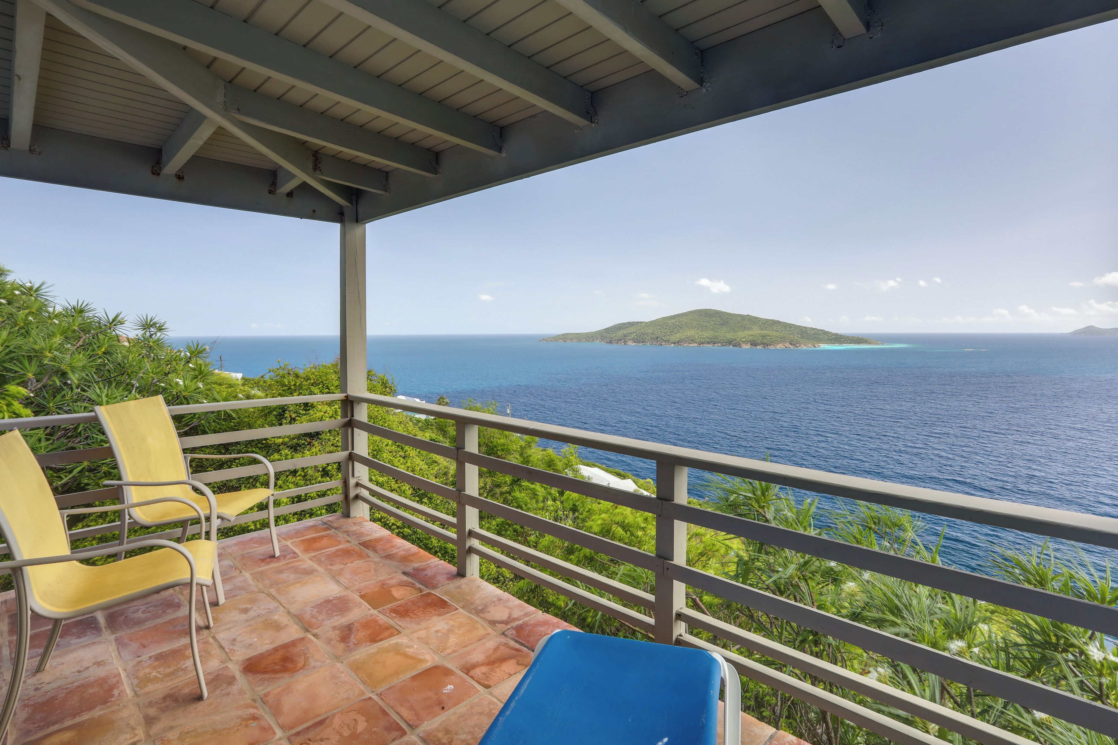 Un espace extérieur aménagé, abrité et doté de chaises et d’une table bleue, offrant une vue panoramique sur l’océan et une île lointaine.