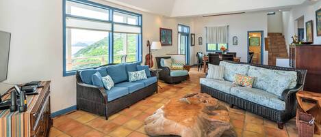 A spacious living area with two sofas, a coffee table, and a TV. Large windows offer a view of the outdoors.