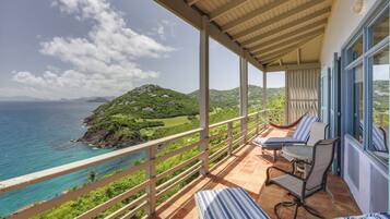 A balcony with a view of the ocean and hills, furnished with chairs and a table.
