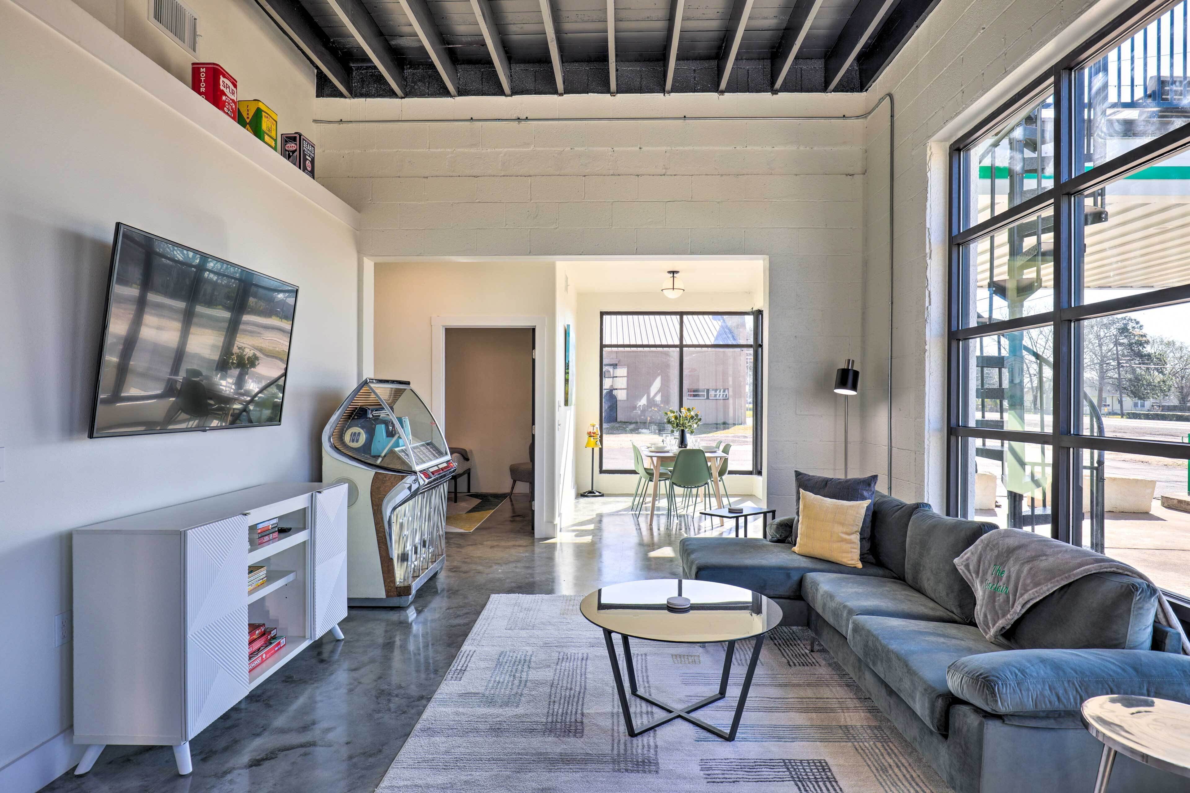 Una sala de estar moderna con un sofá gris, una pequeña mesa redonda y un armario blanco. Una ventana grande ofrece luz natural y vistas al exterior.
