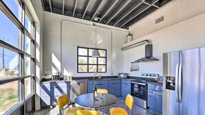 A modern kitchen with a large window, a dining table with chairs, stainless steel appliances, and a view of the outdoors. - Unique Texas Home in Converted Gas Station! (Schulenburg)