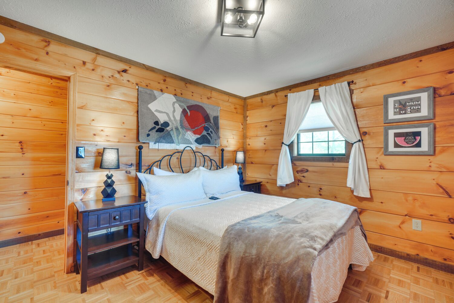 A wooden cabin bedroom with a bed, nightstands, a lamp, a painting, and a window with curtains.