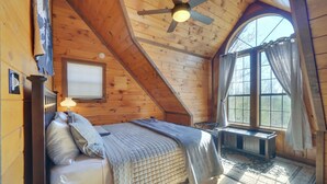 Ein gemütliches Schlafzimmer im Dachgeschoss mit schrägen Wänden, einem Bett mit gesteppter Decke, einem Schreibtisch und einem großen Fenster mit Vorhängen.