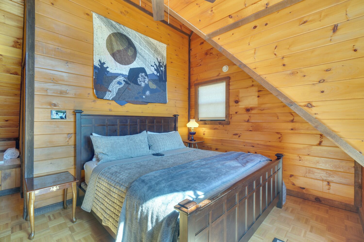 Une chambre dans un chalet en bois, avec un lit, une table de chevet, une lampe fixée au mur et une grande tapisserie.