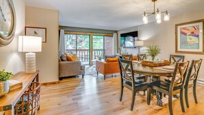 A dining area with a wooden table and chairs, a clock on the wall, and a view of the outdoors. - 1 Block to Main St: Modern Breckenridge Condo! (Breckenridge)