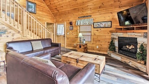 A wooden cabin interior with a leather sofa, a fireplace, a television, and a staircase.