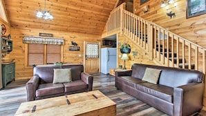 A wooden cabin interior with two leather sofas, a wooden coffee table, a staircase, and a kitchen area.