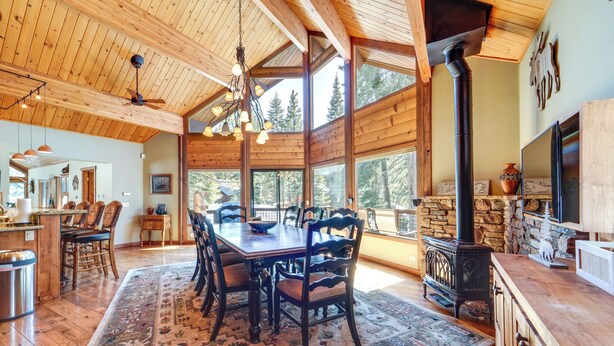 A spacious dining area with a large wooden table and chairs, a stone fireplace, and large windows offering a view of the surrounding forest. - 'one Moose Lodge' Tahoe Donner Vacation Rental! (Truckee)