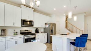 A modern kitchen with white cabinets, stainless steel appliances, a central island with bar stools, and a staircase leading to another level. - 5 Mi to Bethany Beach: Townhome w/ Community Perks (Millville)