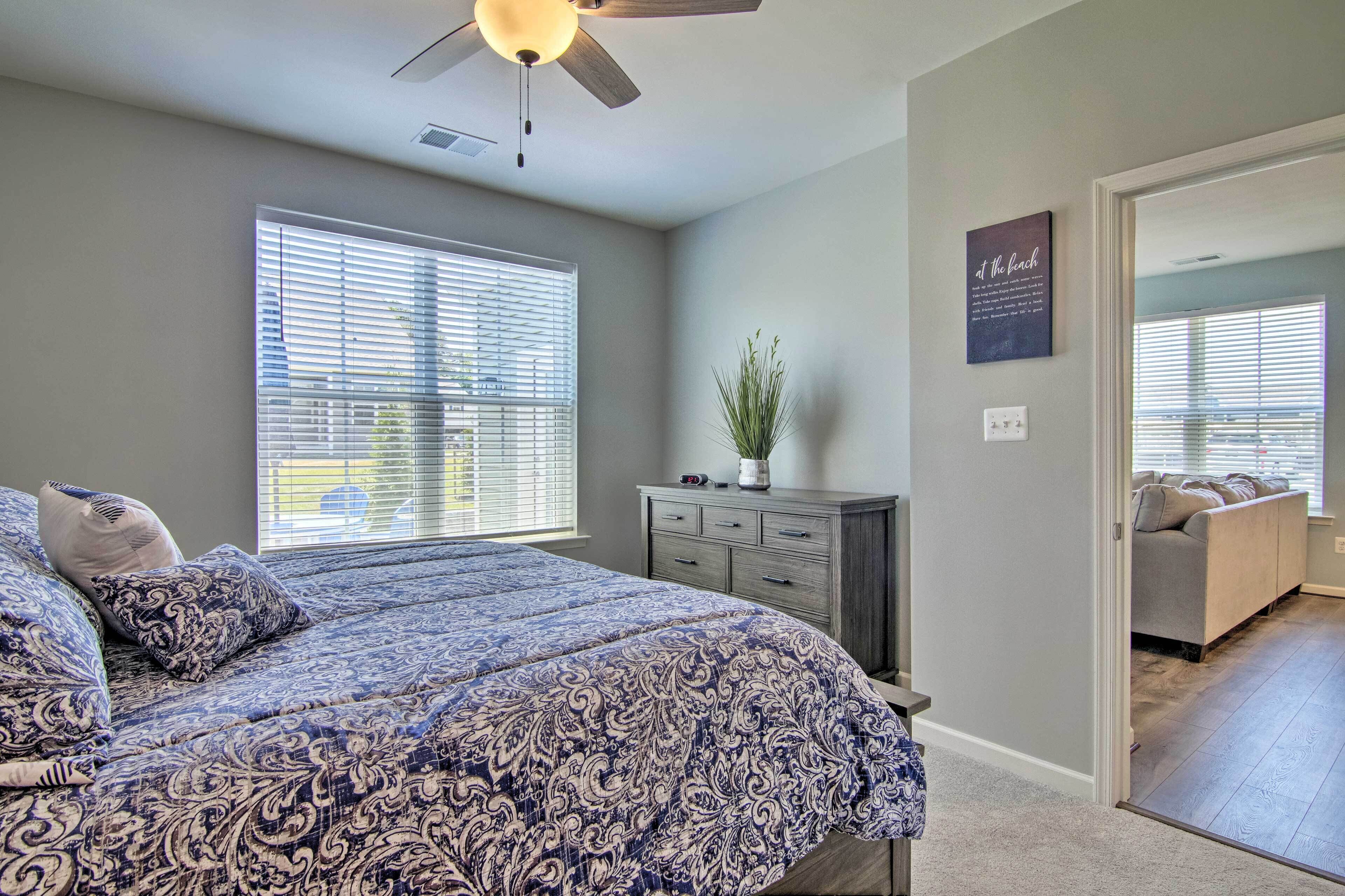 A bedroom with a large bed, a ceiling fan, a dresser, and a window with blinds.