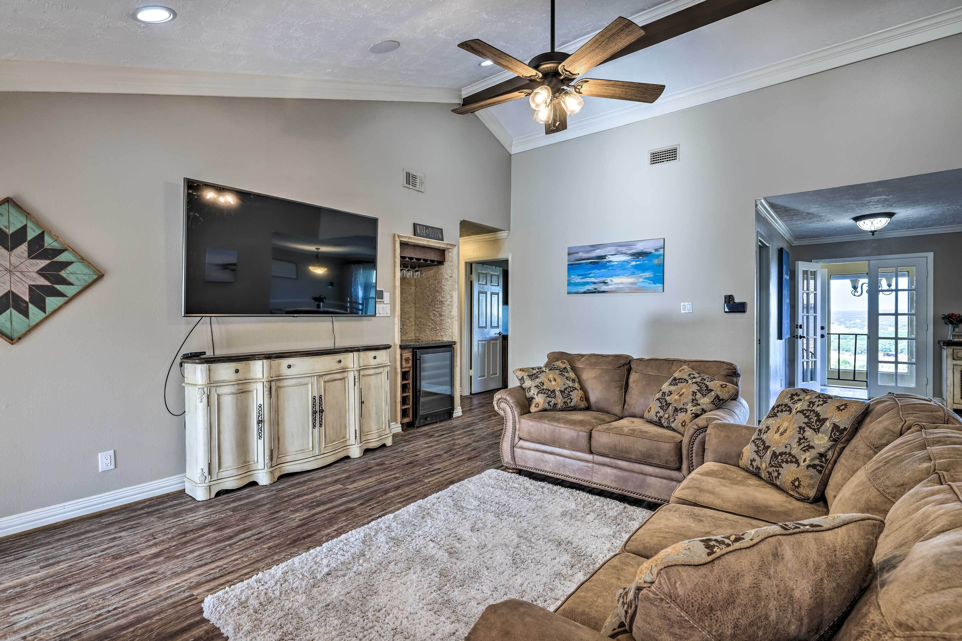 A spacious living room with a large flat-screen TV, a beige sofa set, and a wooden ceiling fan.