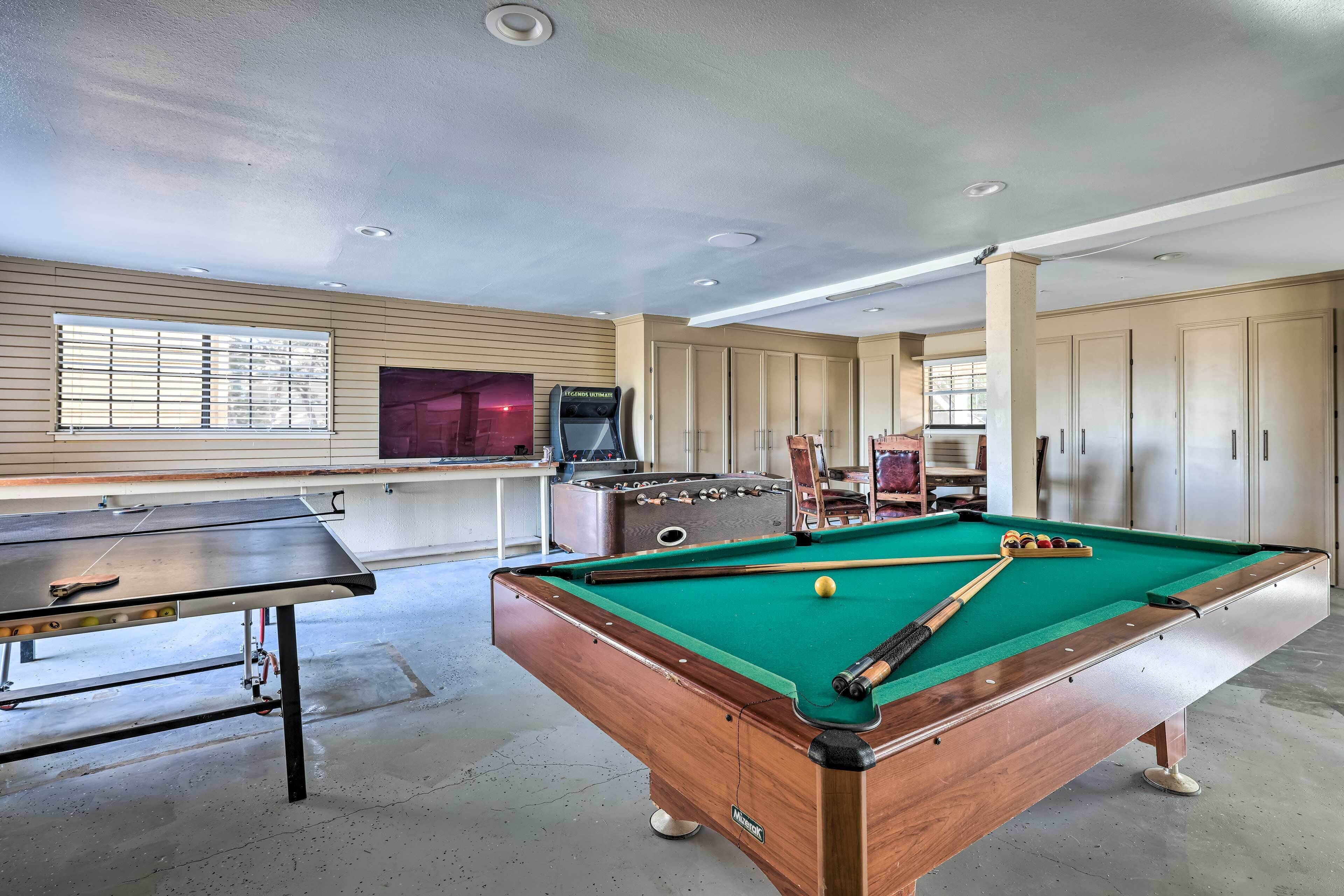 A pool table with a green felt surface, a pool cue, and a cue ball. There is a foosball table and a television in the background.