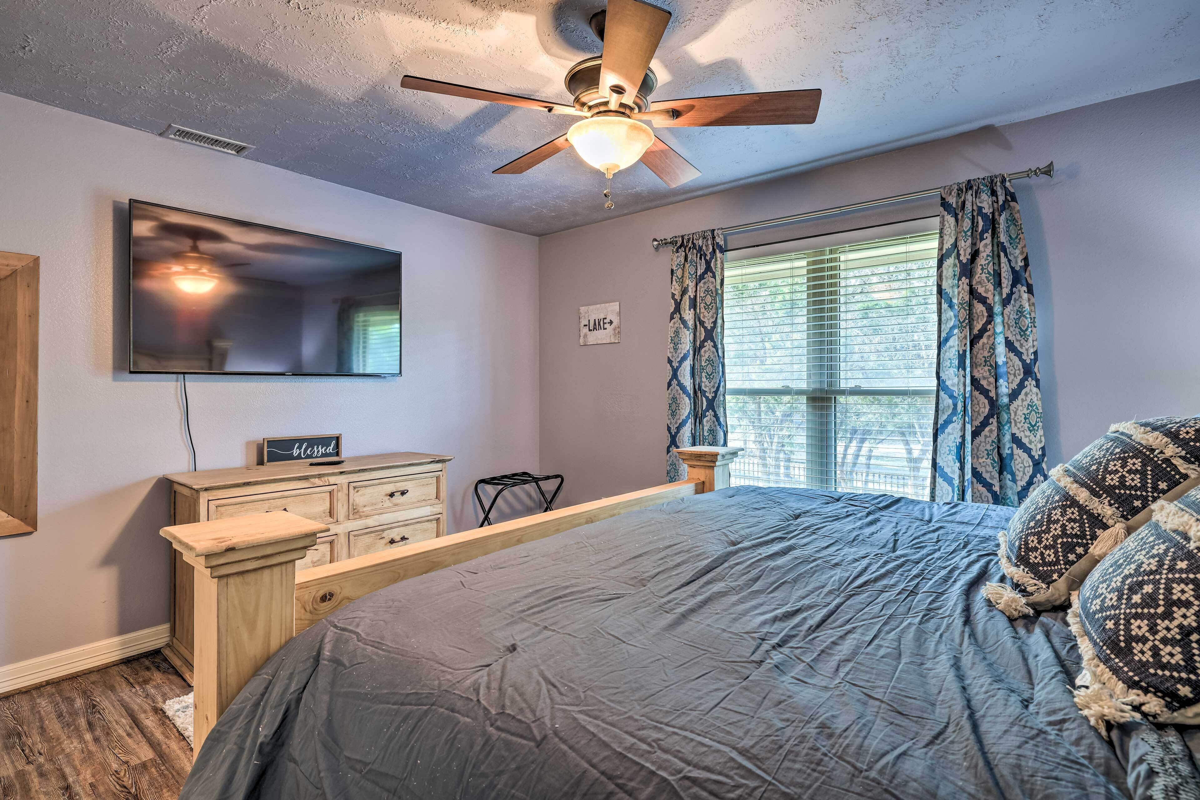 A bedroom with a bed, a ceiling fan, a TV mounted on the wall, a dresser, and a window with patterned curtains.