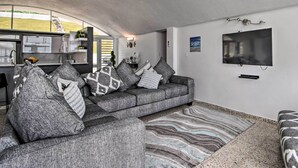 Una sala de estar moderna con un sofá gris, cojines estampados, un televisor de pantalla plana y una cocina visible a través de un arco.