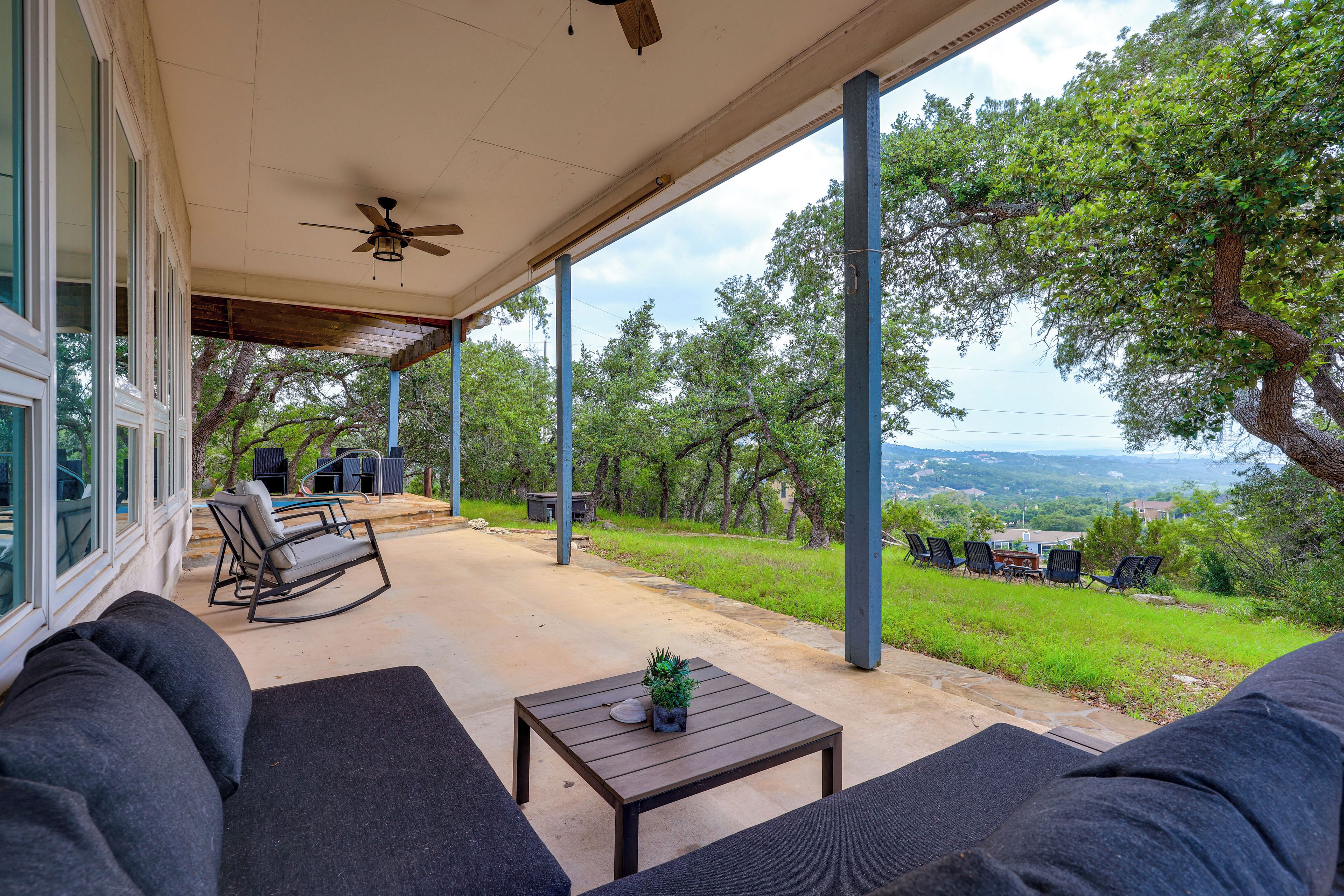 Eine überdachte Terrasse mit Gartenmöbeln, einem Deckenventilator und Blick auf eine Rasenfläche mit Bäumen.
