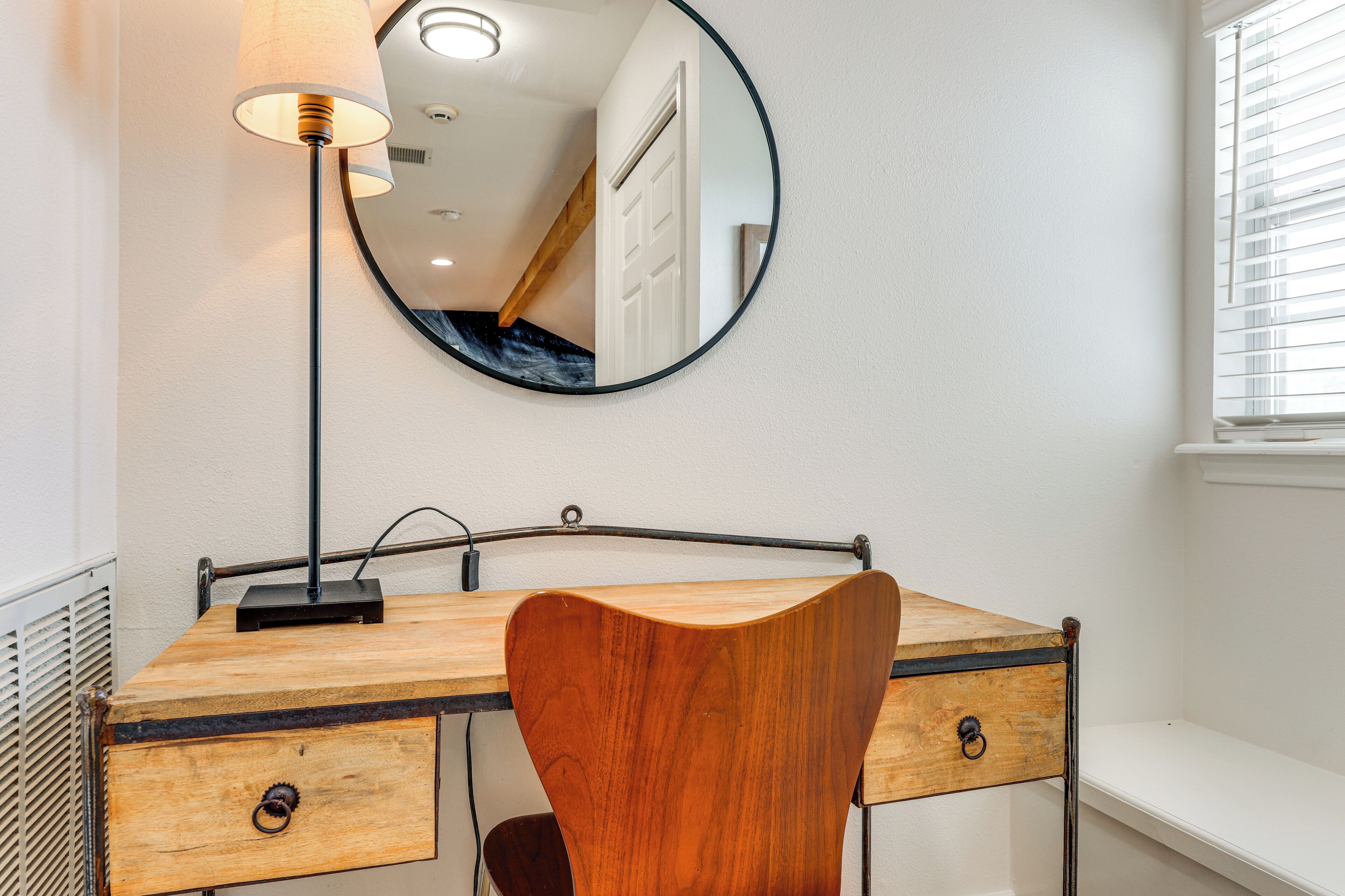 A wooden desk with a mirror, a lamp, and a chair in a room with white walls and a window with blinds.