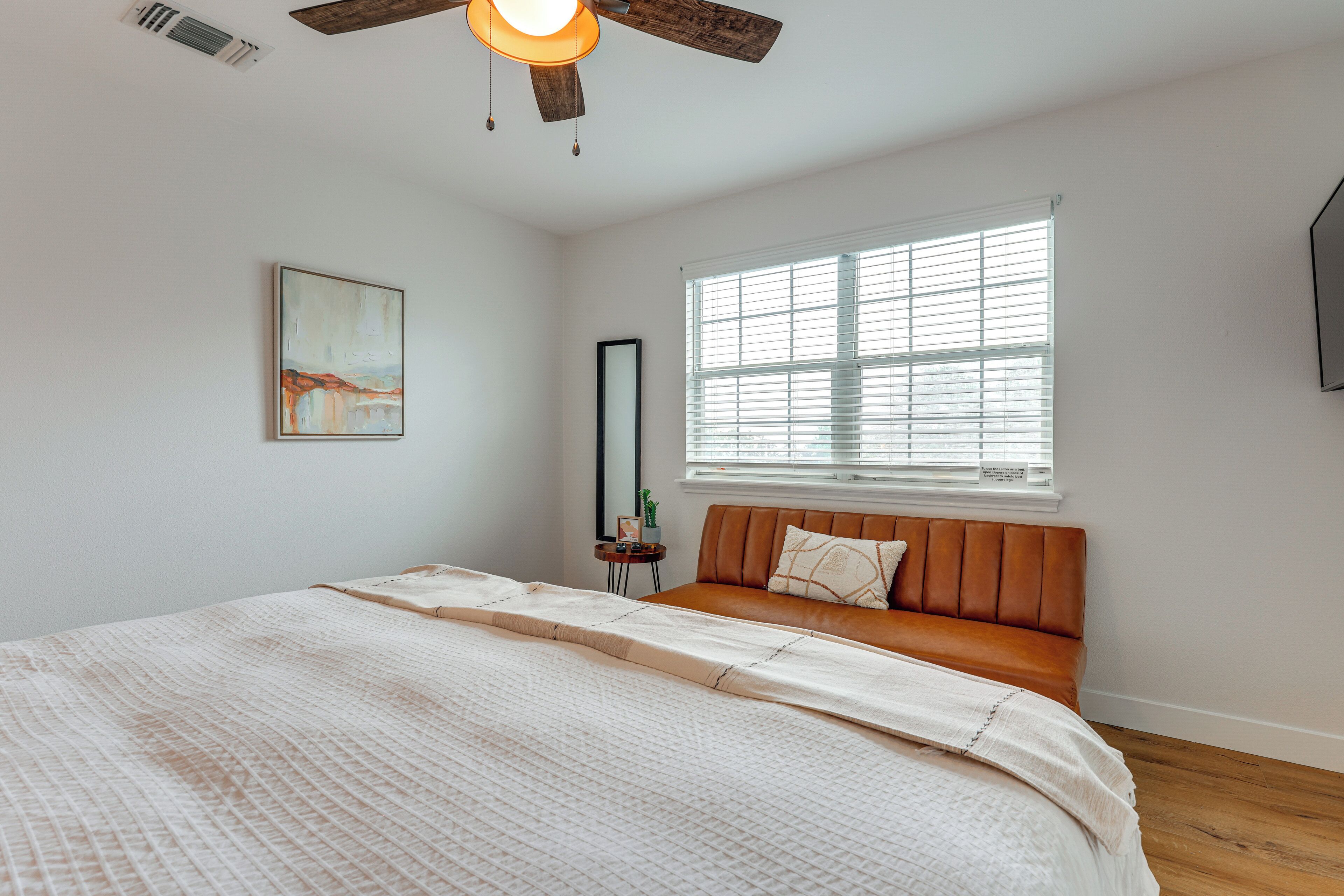 A bedroom with a bed, a window with blinds, a ceiling fan, a wall-mounted mirror, and a small table with a plant.