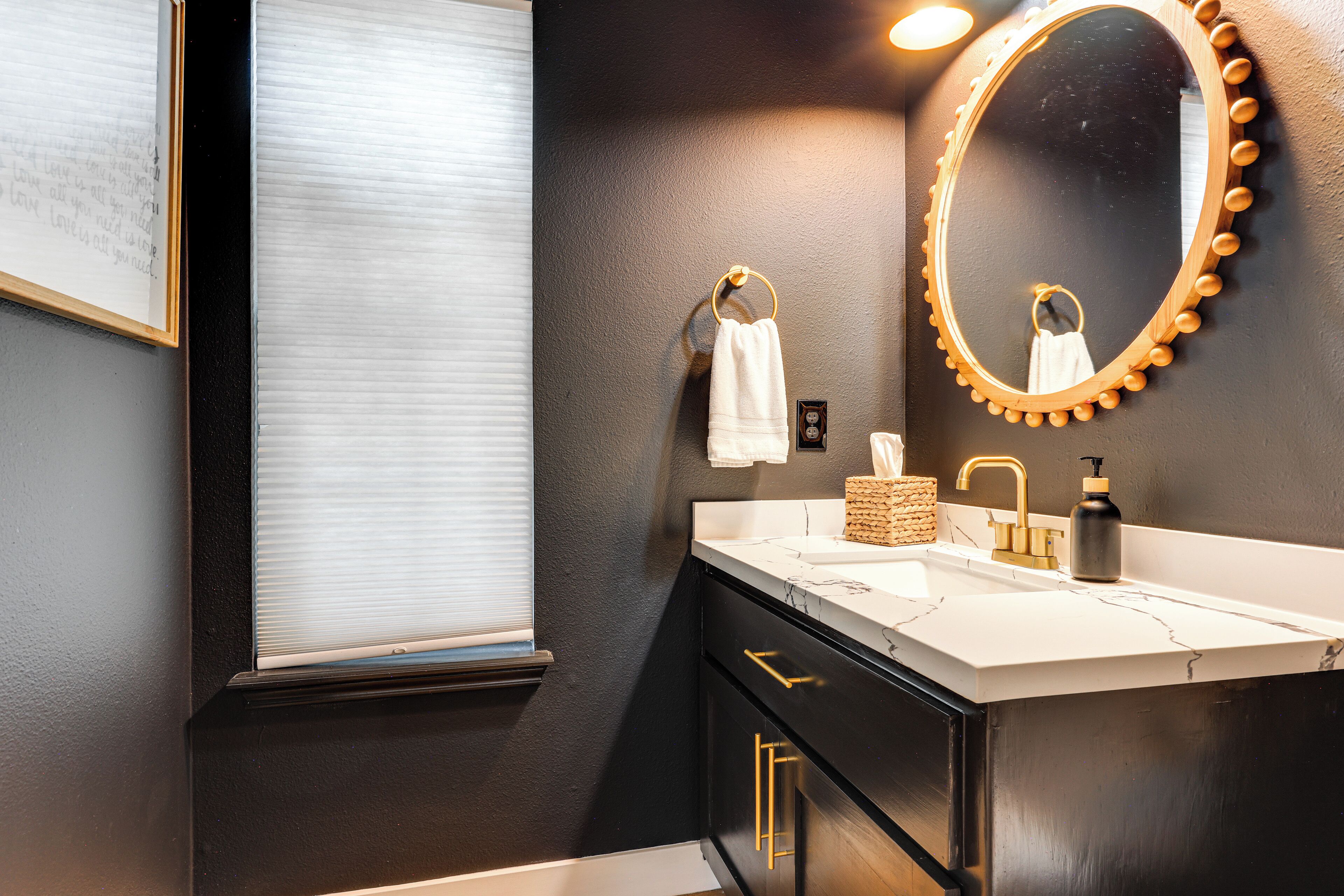 A modern bathroom with a black and white color scheme, featuring a large mirror with a decorative frame, a white sink with gold handles, and a window with blinds.