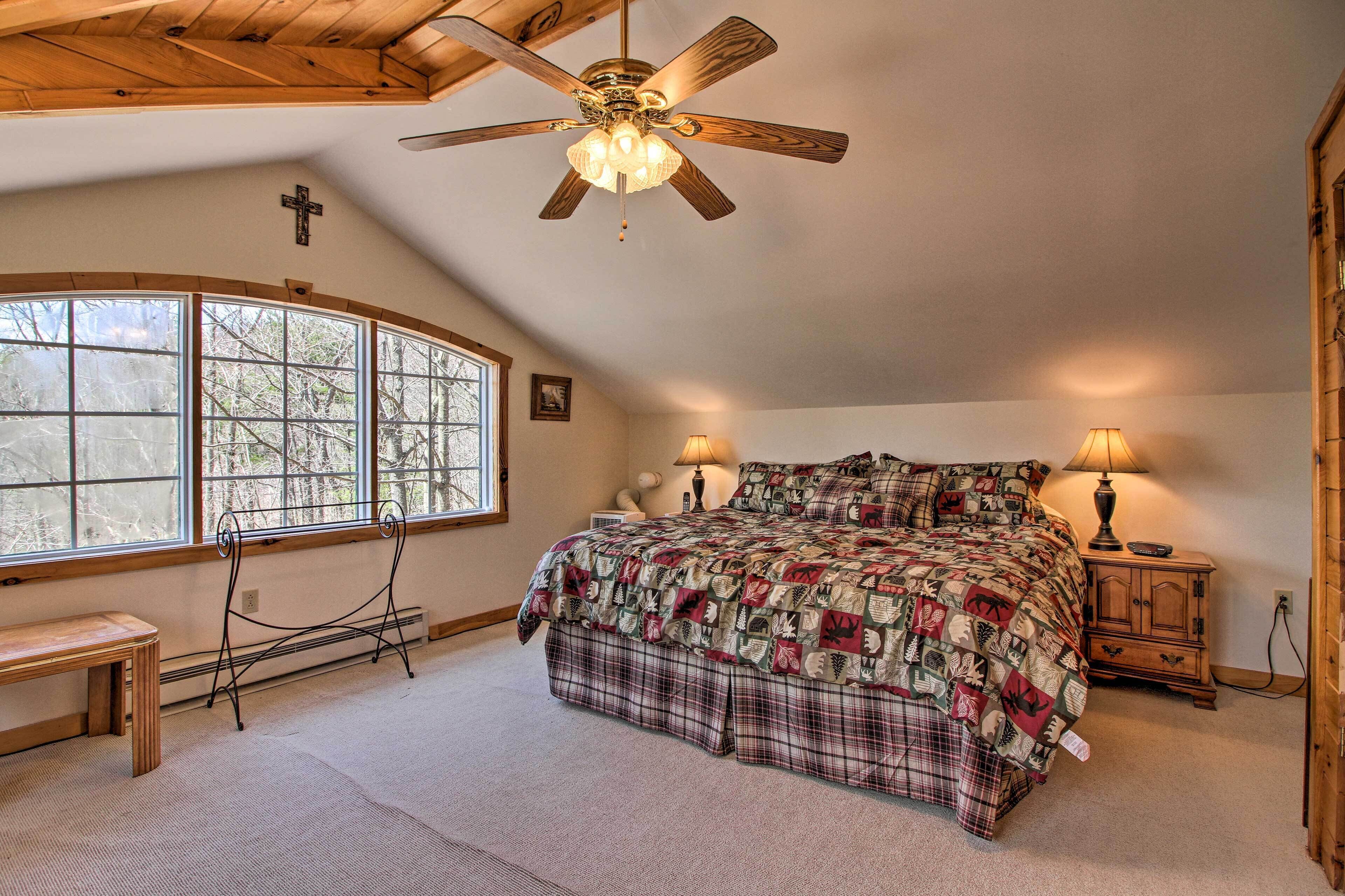 A bedroom with a large bed, a ceiling fan, a bench, a window, and a nightstand with a lamp.