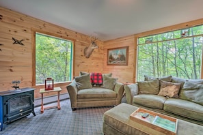 A cozy cabin interior with a fireplace, a sofa, a loveseat, and a large window offering a forest view.