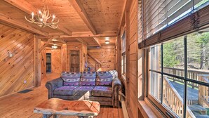 A log cabin interior with a wooden ceiling, a chandelier, a sofa with patterned cushions, and a large window overlooking a deck.
