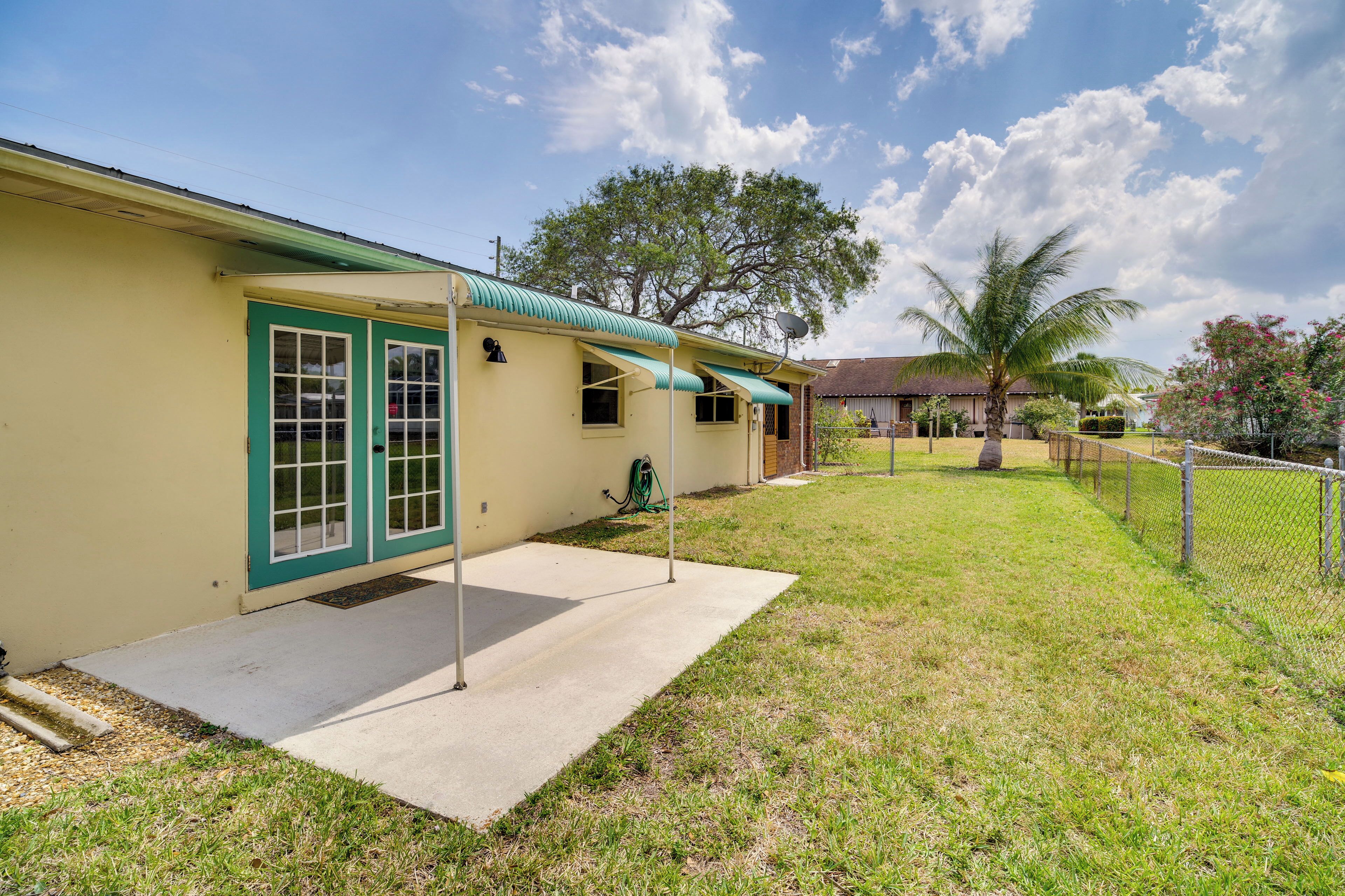 A single-story house with a green door and windows, a covered porch, and a fenced yard with a palm tree.