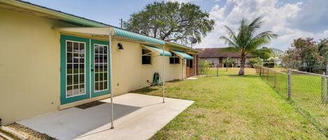A single-story house with a green door and windows, a covered porch, and a fenced yard with a palm tree.