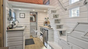 A compact kitchen with white cabinetry, a small refrigerator, and a sink. There is a window on the right side and a door on the left. - Unique Stay: 'tiny Everest' San Antonio Home! (San Antonio)