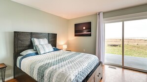 Un dormitorio con una cama grande, una mesita de noche, un cuadro en la pared y vista a la playa a través de un ventanal corredero.