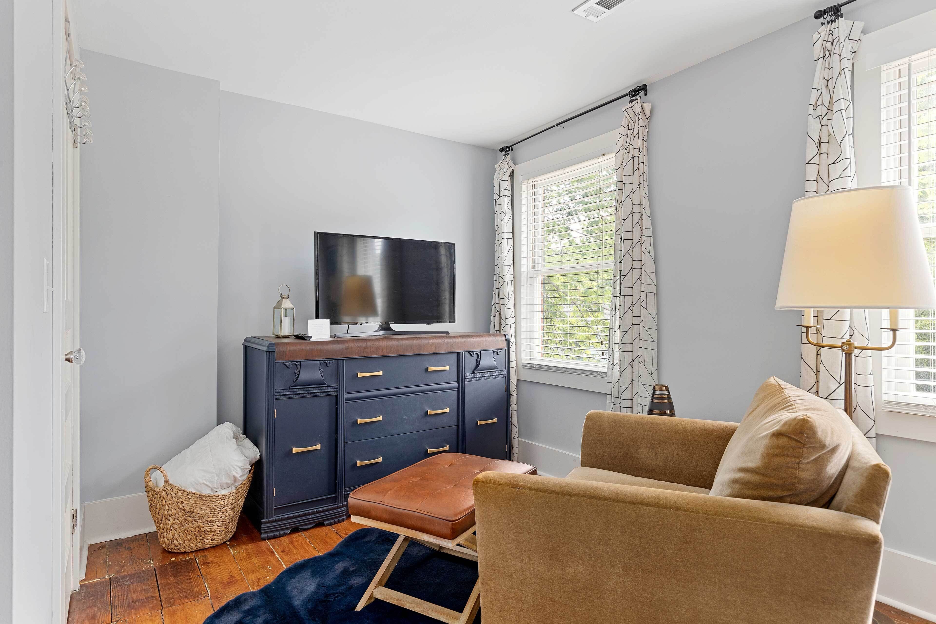 Una habitación con un aparador azul oscuro, un televisor encima, un sillón beige, una ventana con cortinas estampadas y una lámpara.
