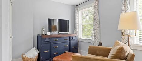 Una habitación con un aparador azul oscuro, un televisor encima, un sillón beige, una ventana con cortinas estampadas y una lámpara.