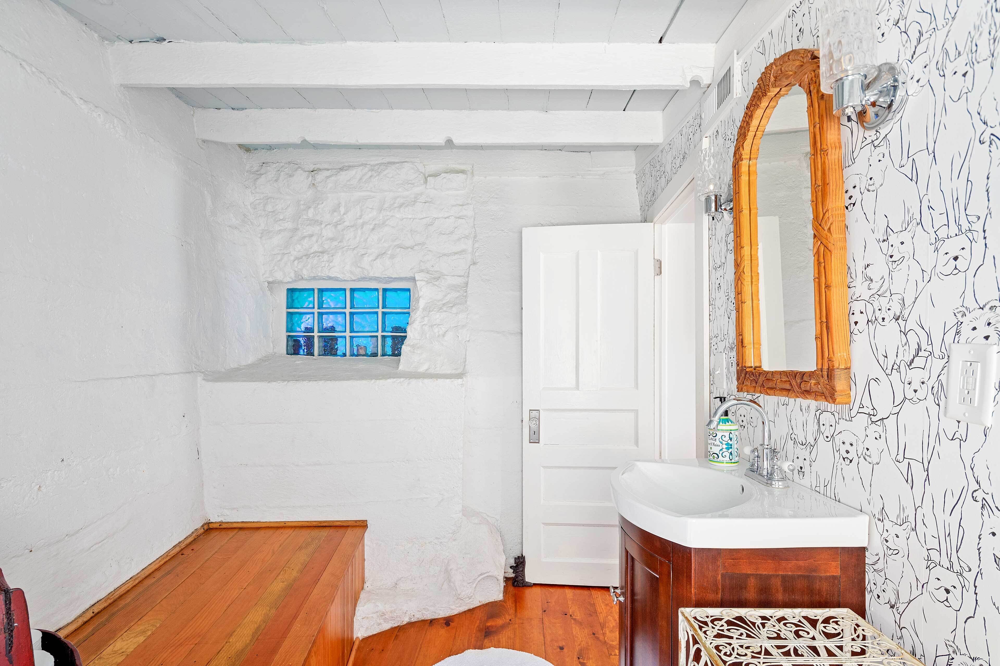 Ein Badezimmer mit weißer Steinwand, Holzboden, einer weißen Tür, einem weißen Waschbecken mit Spiegel und einem Fenster mit blau getöntem Glas.