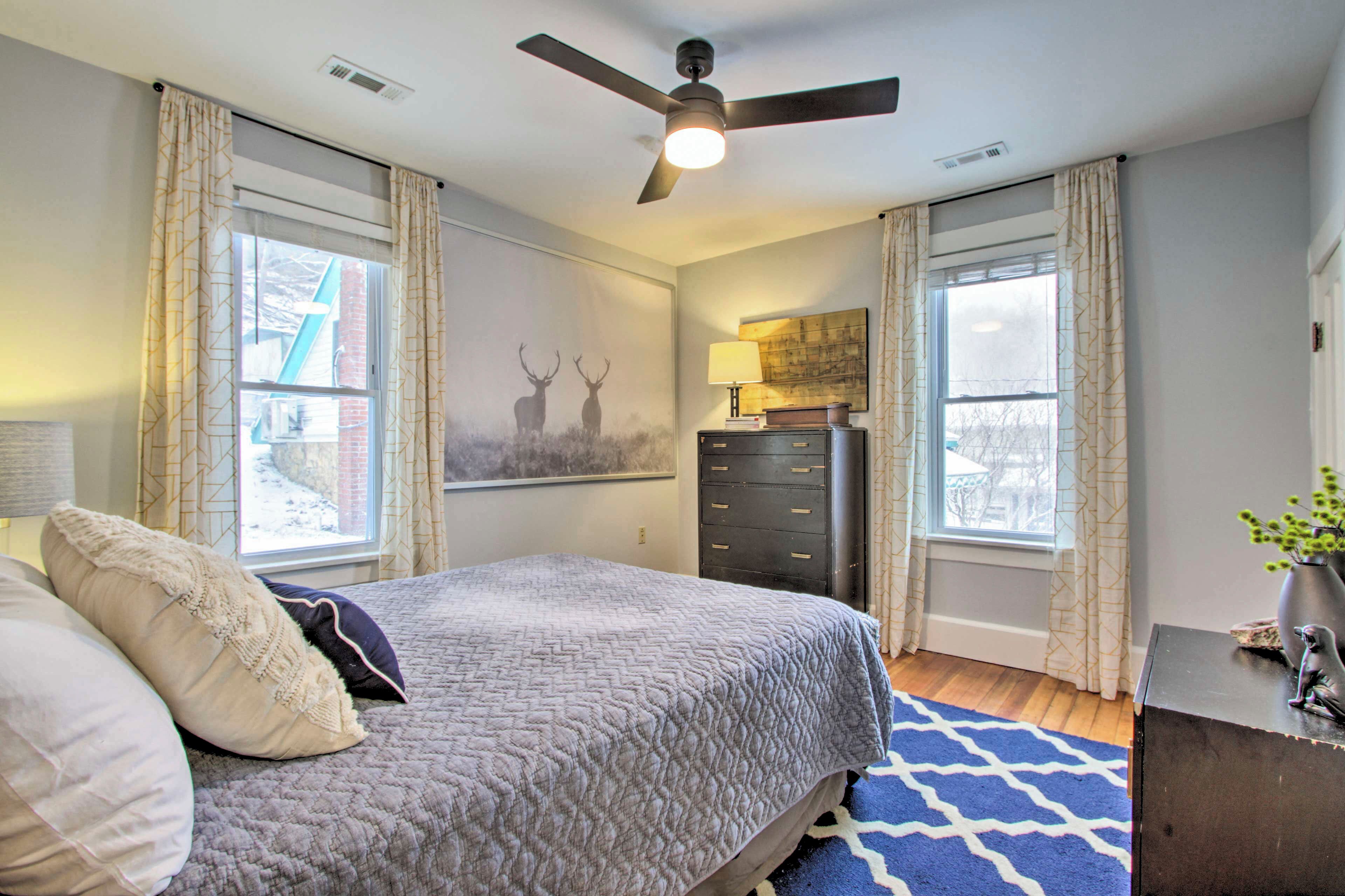 Une chambre à coucher avec un lit, un ventilateur de plafond, une commode et une fenêtre avec des rideaux.