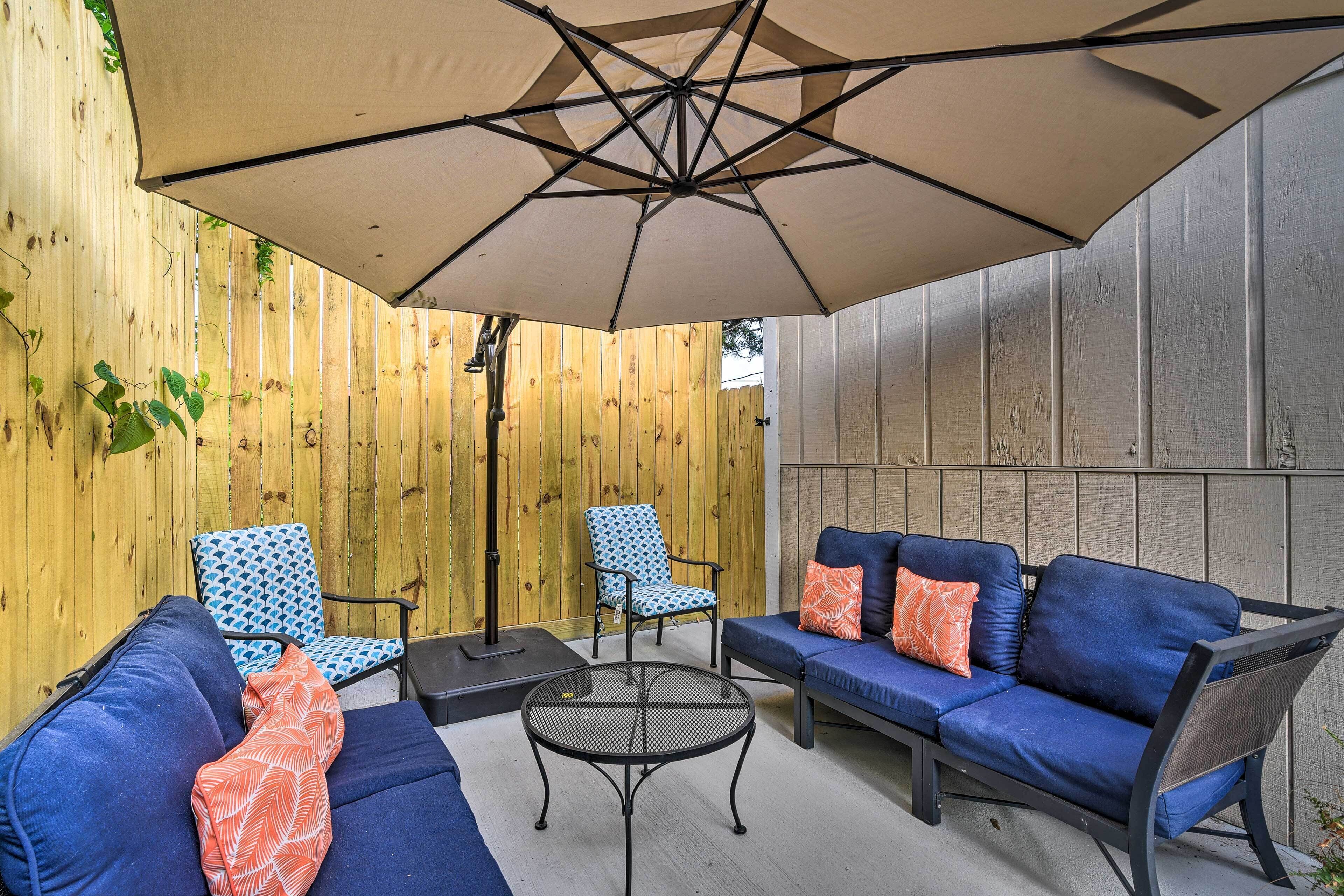 Ein Patio mit einer blauen Sofagarnitur, zwei gemusterten Stühlen, einem runden Tisch und einem großen Sonnenschirm, der Schatten spendet.