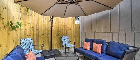 A patio area with a blue sofa set, two patterned chairs, a round table, and a large umbrella providing shade.