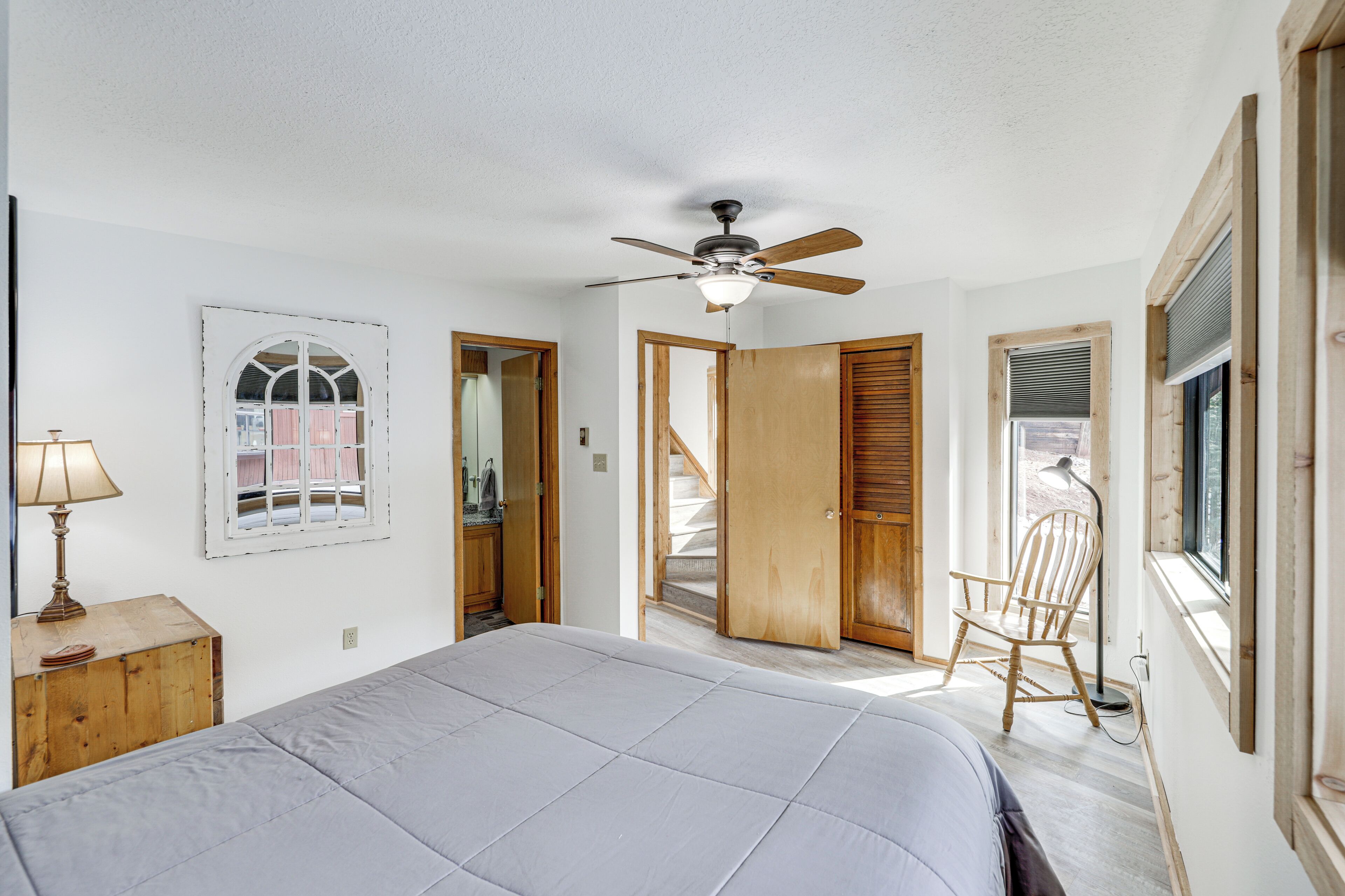 A bedroom with a bed, a ceiling fan, a window, a chair, and a wooden door.