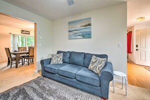 Un salon avec un canapé bleu, des coussins à motifs, un tapis à longues mèches gris, un tableau au mur et, au fond, un coin repas.