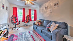 A modern living room with a grey sofa, red curtains, a ceiling fan, and gaming chairs.
