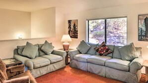 A living room with two grey sofas, a small table with a lamp, a chair, and a large window with a view of trees.