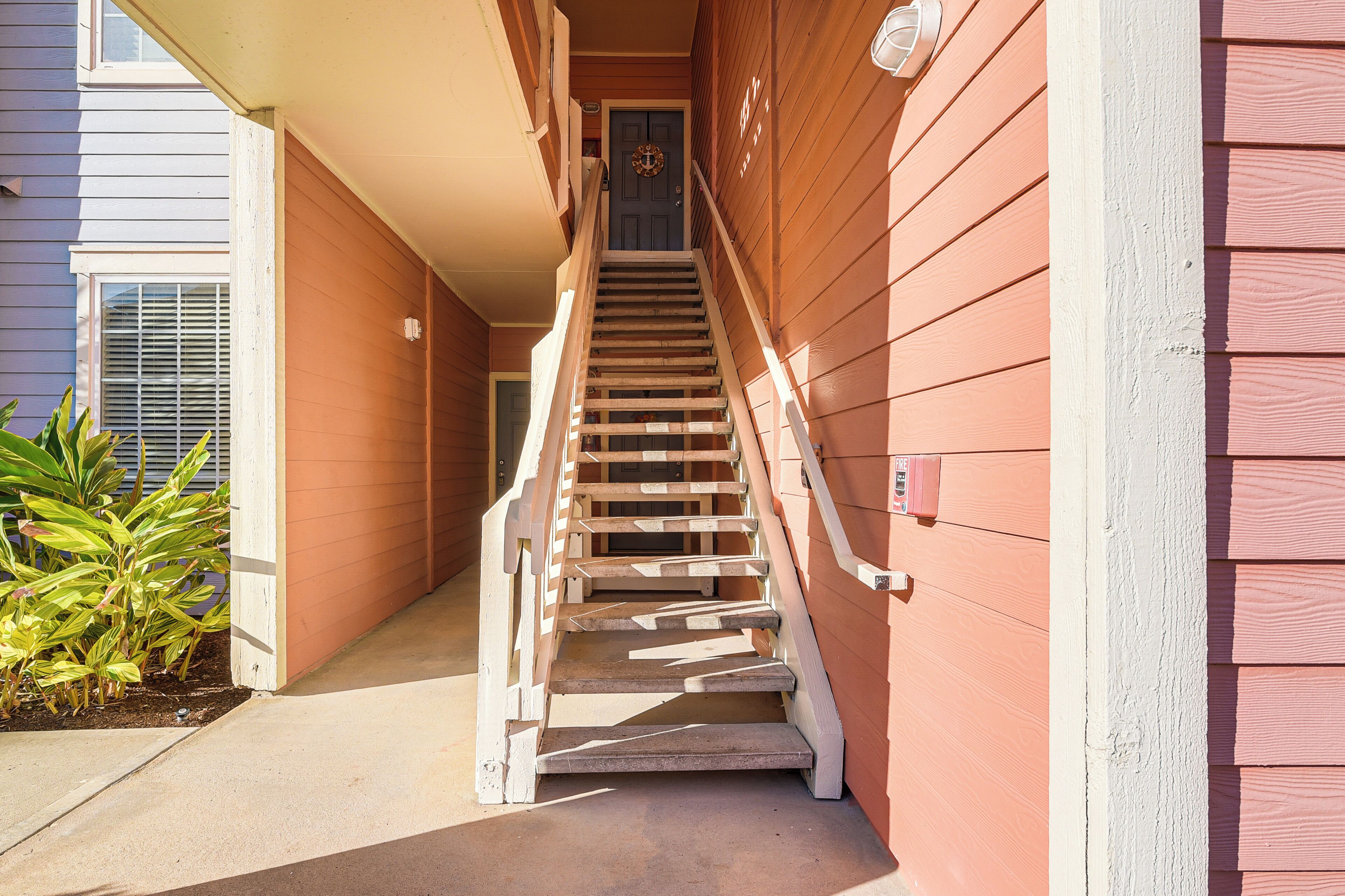 A wooden staircase leading to a door with a wreath, flanked by a plant and a building with a window and a door.