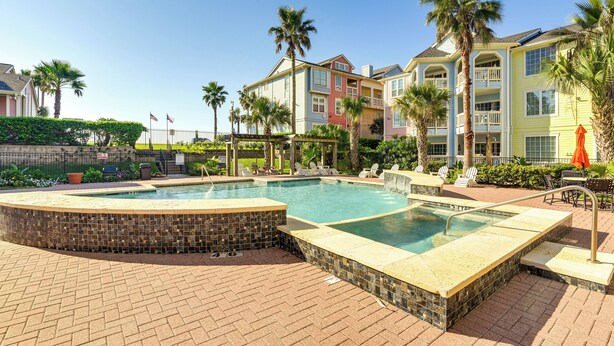 Une piscine avec une terrasse carrelée, entourée de palmiers et bordée d’un immeuble à plusieurs étages doté de balcons.