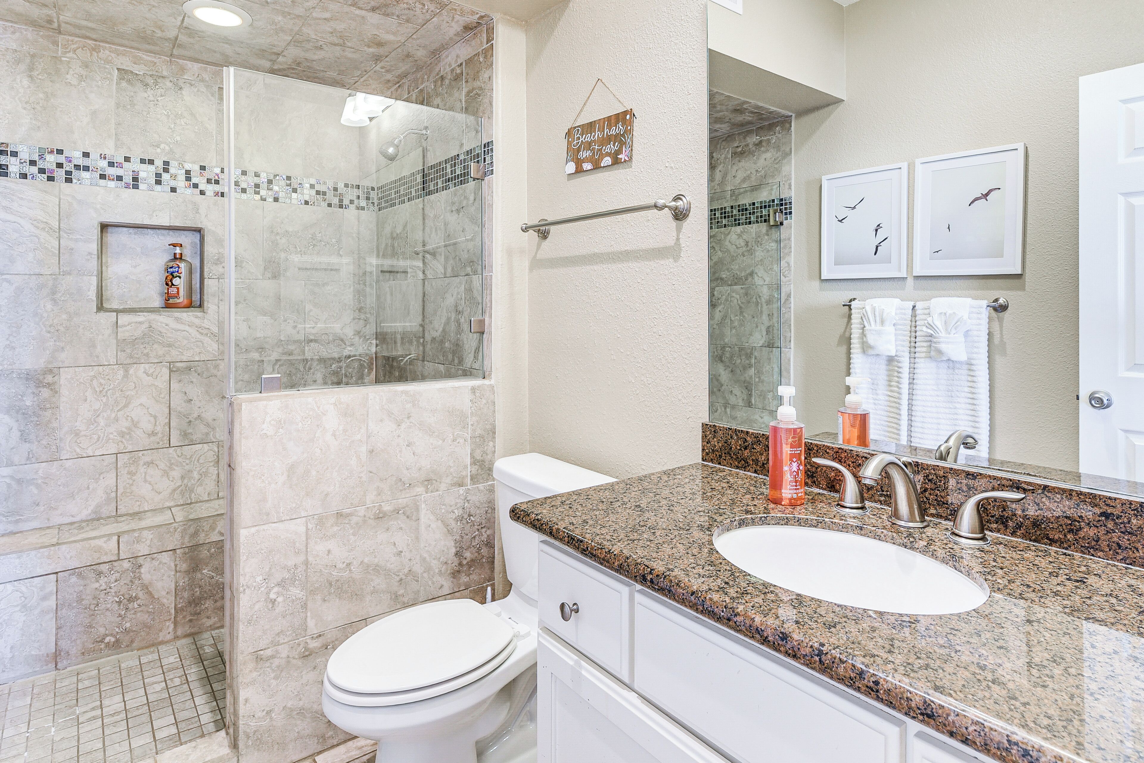 A bathroom with a glass-enclosed shower, a white toilet, a granite countertop with a sink, and two framed pictures on the wall.
