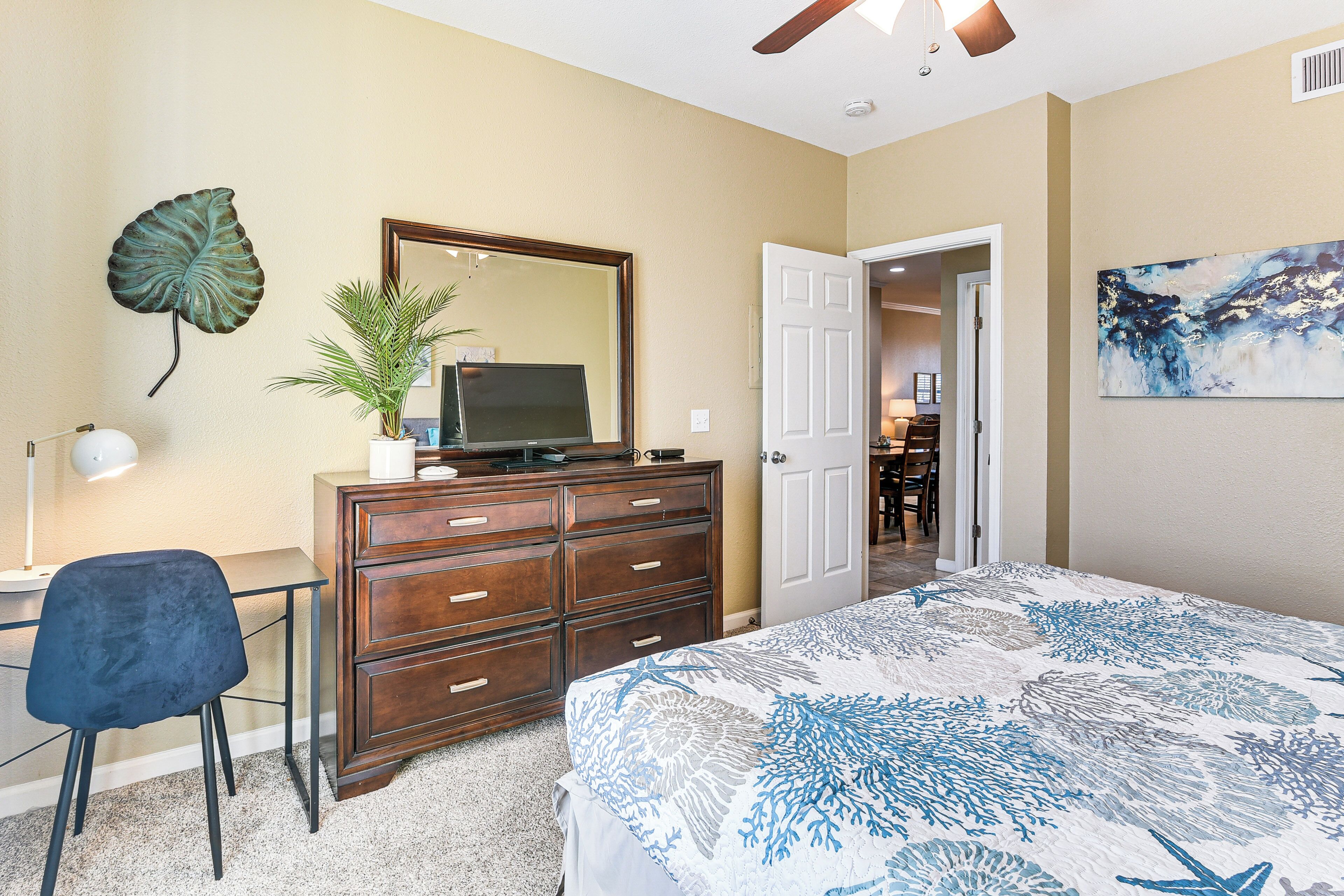 A bedroom with a bed, a dresser, a desk, a chair, a plant, a mirror, and a door leading to another room.