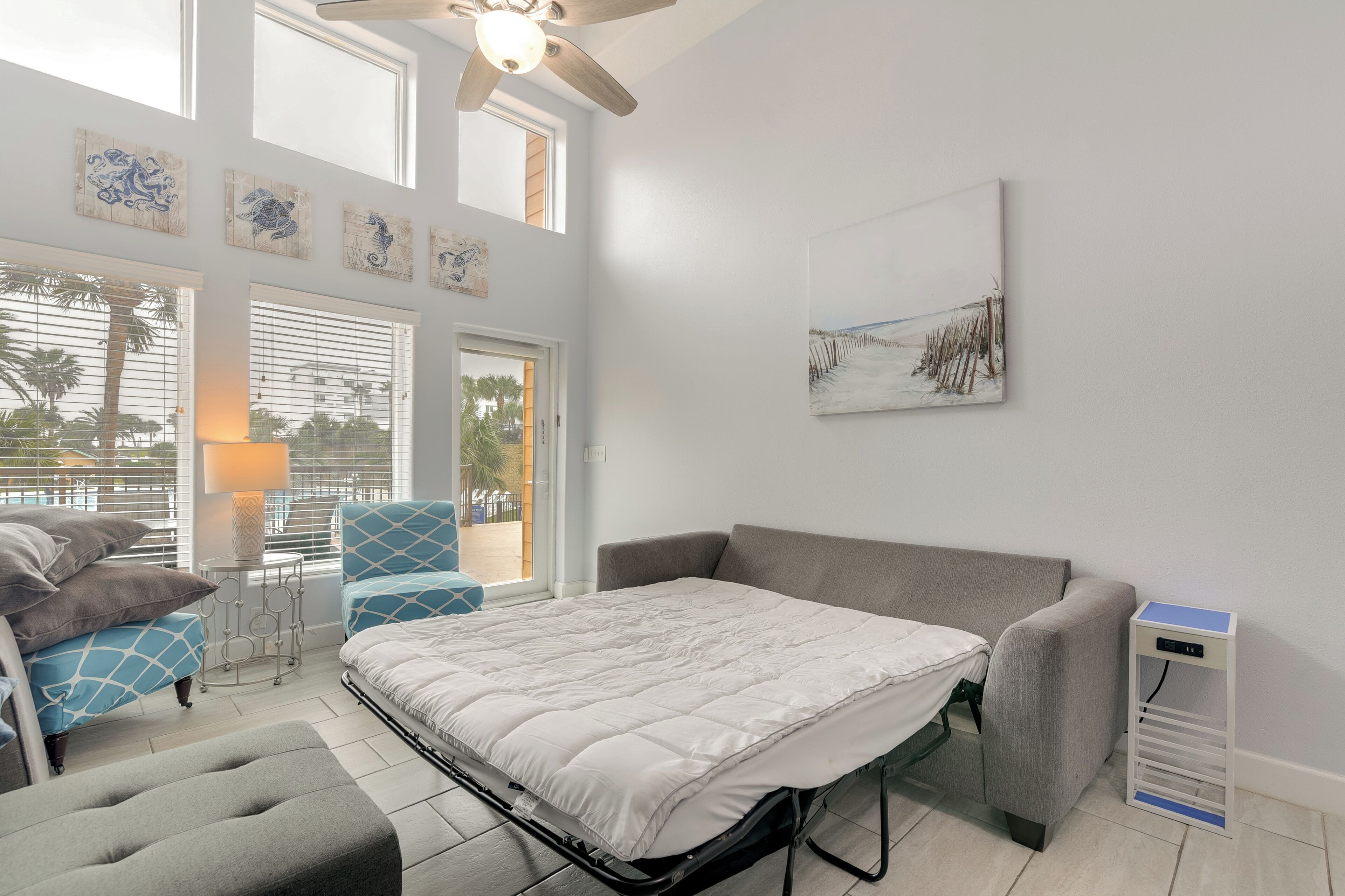A room with a bed, a grey sofa, a blue armchair, and a small table with a lamp. There are windows with blinds, a ceiling fan, and artwork on the walls.