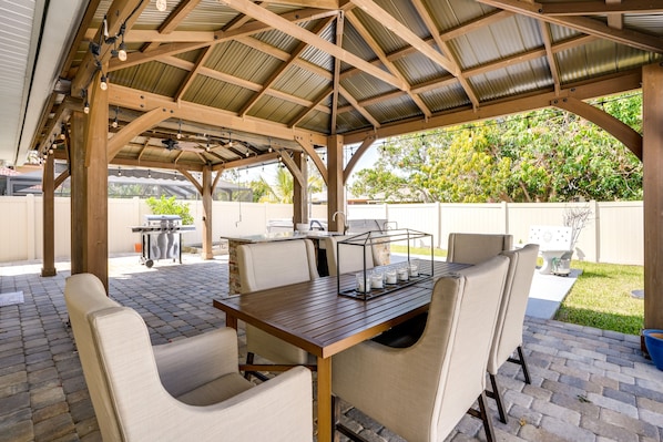 A covered patio with a wooden table and chairs, a grill, and a fenced backyard.