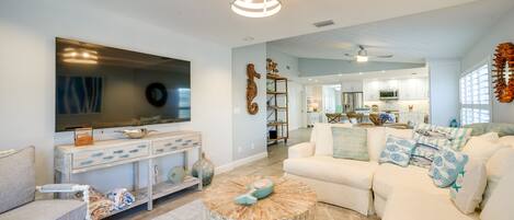 A modern living room with a large flat-screen TV, a round coffee table, and a white sofa with patterned cushions.