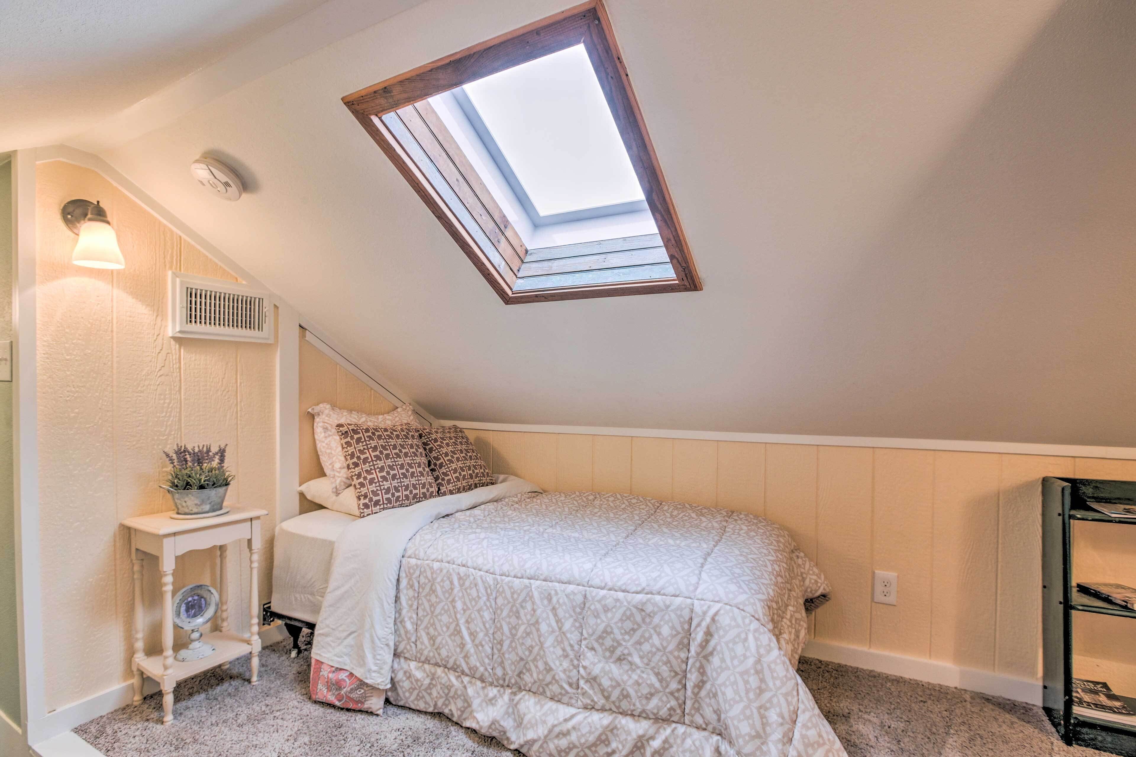 A bedroom with a skylight, a bedside table with a lamp, a bed with patterned bedding, and a small shelf with decorative items.