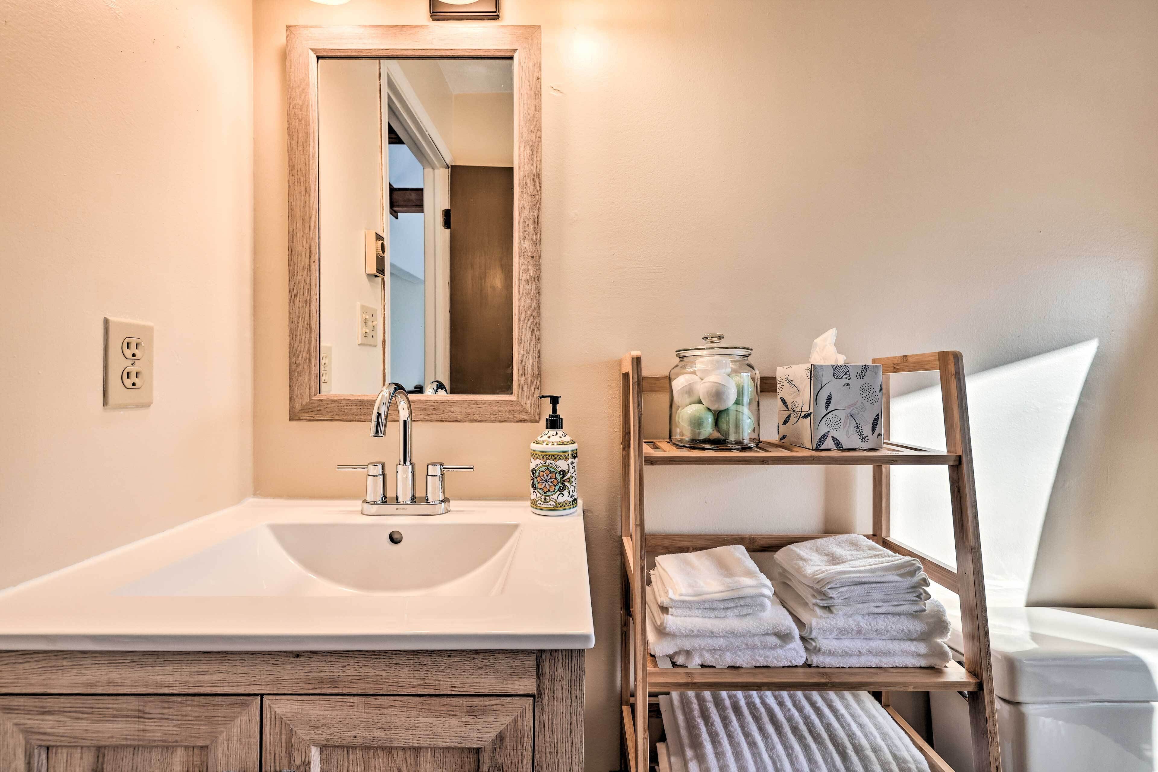 Une salle de bain avec un grand lavabo, un miroir, une étagère en bois avec des serviettes et un petit meuble contenant des articles de toilette.